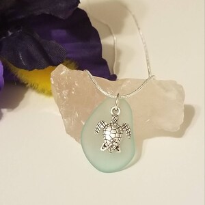 May include: A silver turtle charm pendant hangs from a silver chain on a light blue sea glass pendant. The pendant is resting on a pink and white rock.