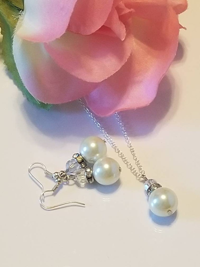 May include: A pearl necklace and earrings set with a silver chain and white pearls. The necklace has a single pearl pendant and the earrings have two pearls each.