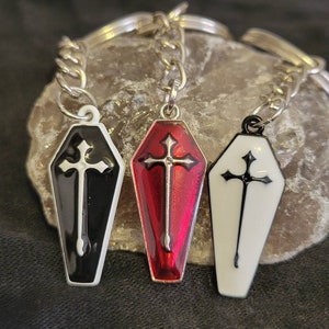 May include: Three coffin-shaped keychains with crosses. One is black with a white cross, one is red with a white cross, and one is white with a black cross.