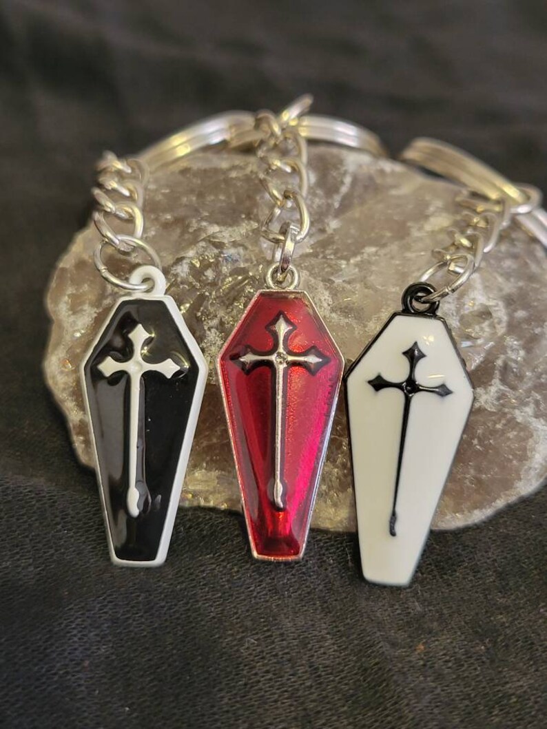 May include: Three coffin-shaped keychains with crosses. One is black with a white cross, one is red with a white cross, and one is white with a black cross.