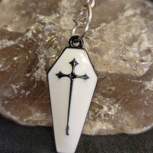 May include: A black and white coffin-shaped keychain with a silver chain. The coffin has a black cross with a white background.