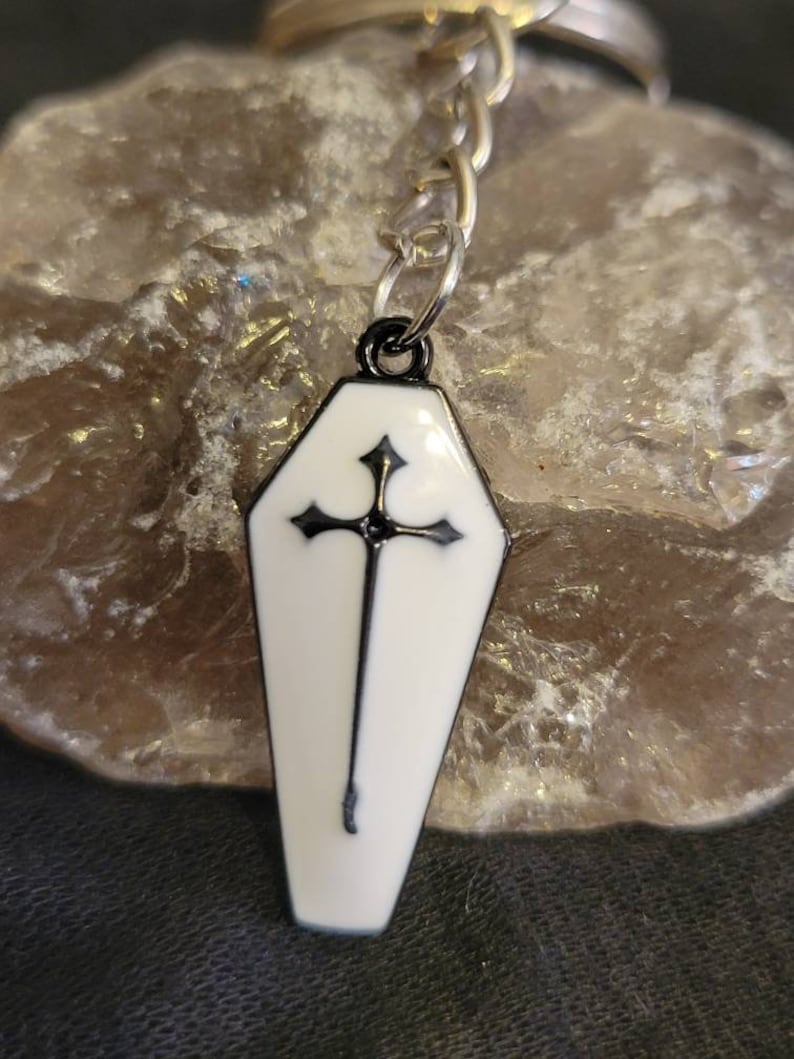 May include: A black and white coffin-shaped keychain with a silver chain. The coffin has a black cross with a white background.
