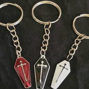 May include: Three silver keychains with coffin-shaped charms. The charms are red, black, and white with a silver cross on each.