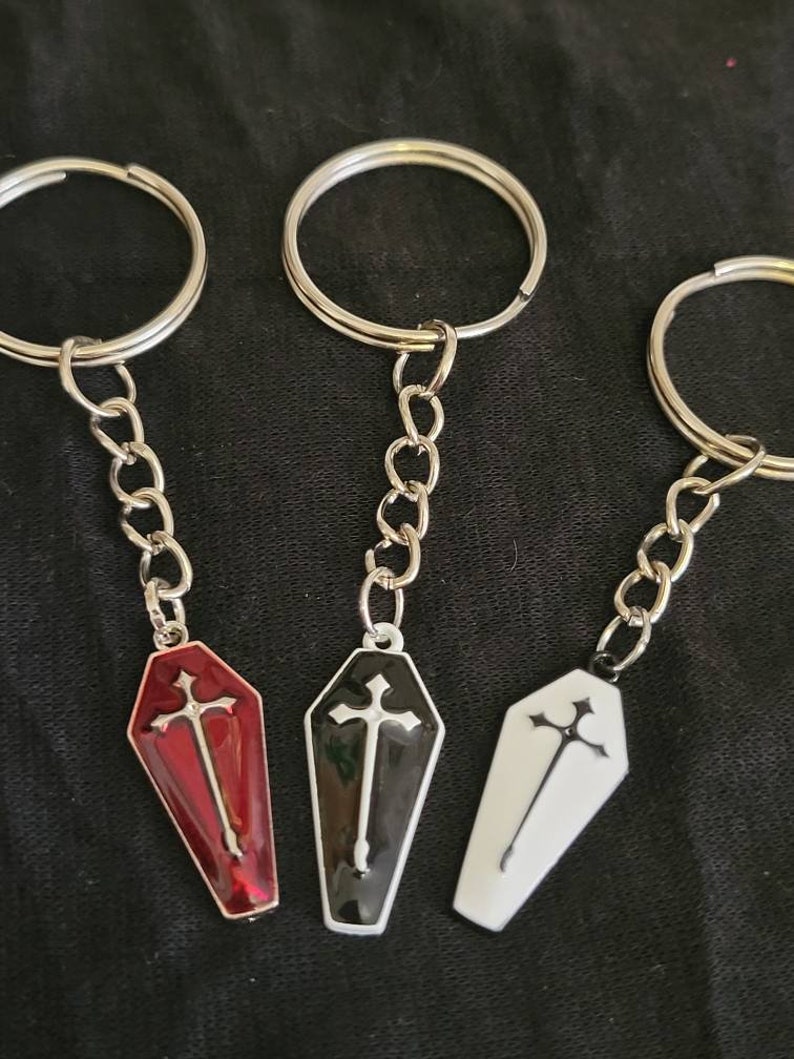 May include: Three silver keychains with coffin-shaped charms. The charms are red, black, and white with a silver cross on each.