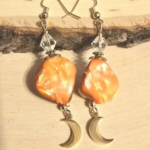 May include: A pair of dangle earrings featuring irregular, polished orange shell beads suspended from silver-toned hooks. Each earring includes a clear, faceted crystal bead above the shell and a small, smooth silver-toned crescent moon charm below.
