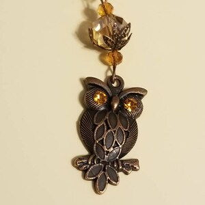 May include: A bronze owl charm with gold eyes, perched on a branch, hangs from a necklace with amber glass beads.