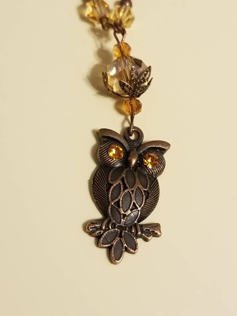 May include: A bronze owl charm with gold eyes, perched on a branch, hangs from a necklace with amber glass beads.