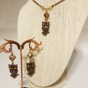 May include: A copper owl necklace and earrings set with amber glass beads. The necklace features a delicate chain and a small owl pendant. The earrings are dangle style with a larger owl charm.