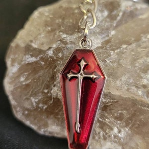 May include: A silver keychain with a red coffin-shaped charm. The charm has a silver cross inside.