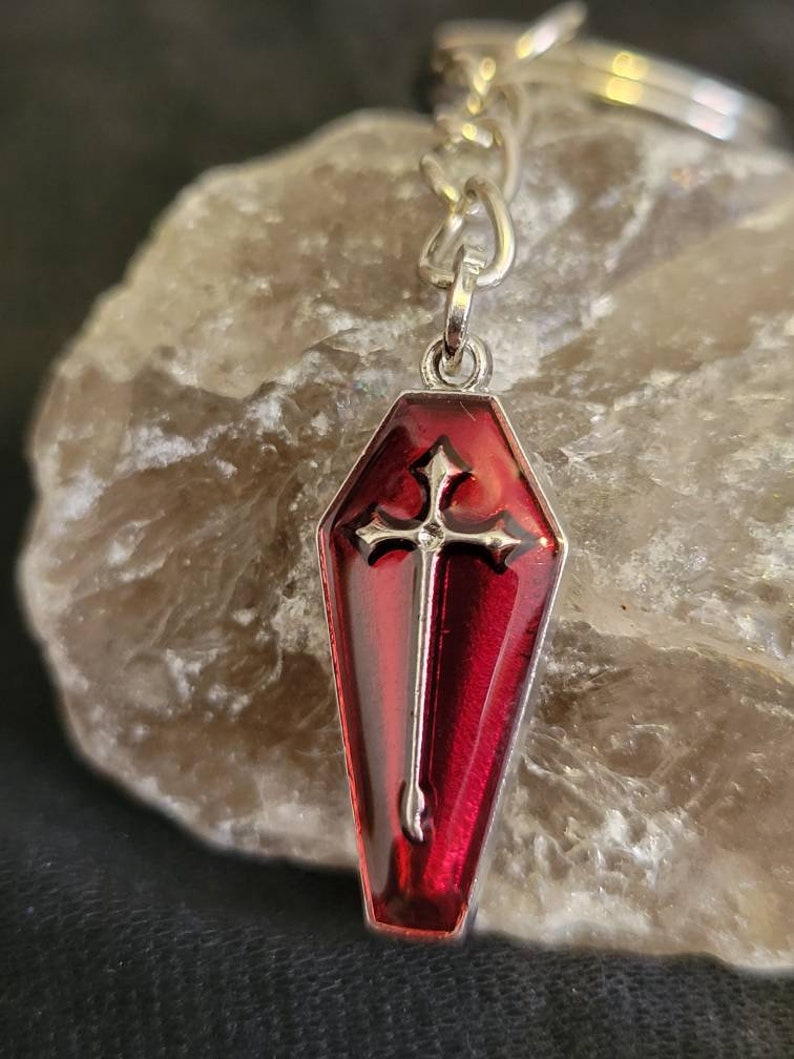 May include: A silver keychain with a red coffin-shaped charm. The charm has a silver cross inside.