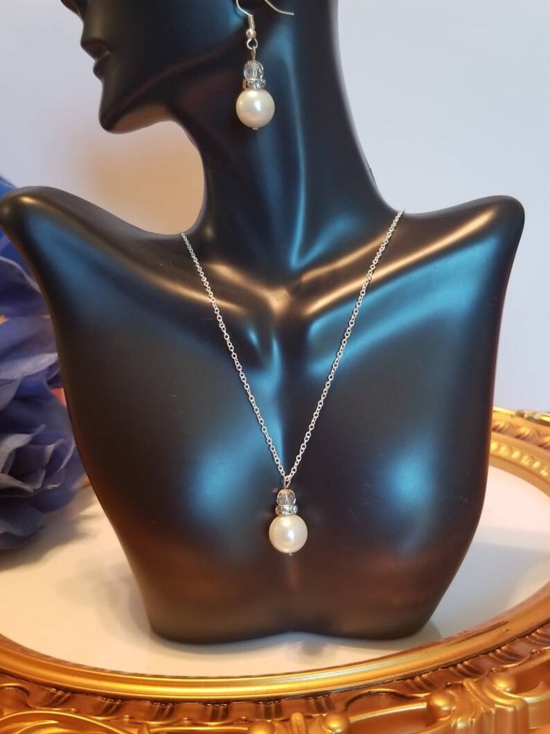 May include: A silver necklace and earrings set with a pearl and crystal pendant. The necklace has a delicate silver chain and the earrings are drop style.