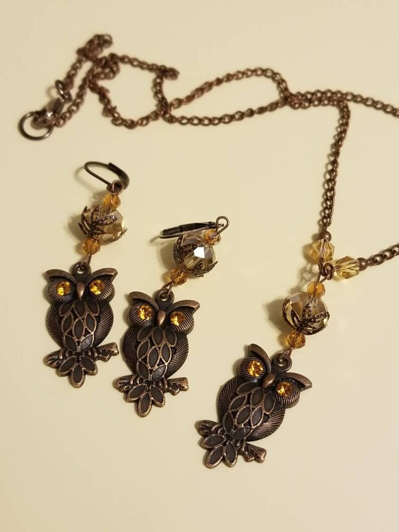 May include: A set of owl-shaped earrings and a necklace with amber-colored crystal beads and bronze-colored metal accents.