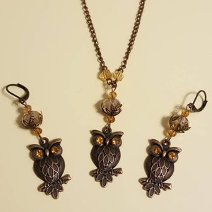 May include: A set of owl jewelry featuring a necklace and earrings. The necklace has a bronze chain with a clear crystal pendant and amber-colored beads. The earrings are bronze with amber-colored crystal accents and owl charms.
