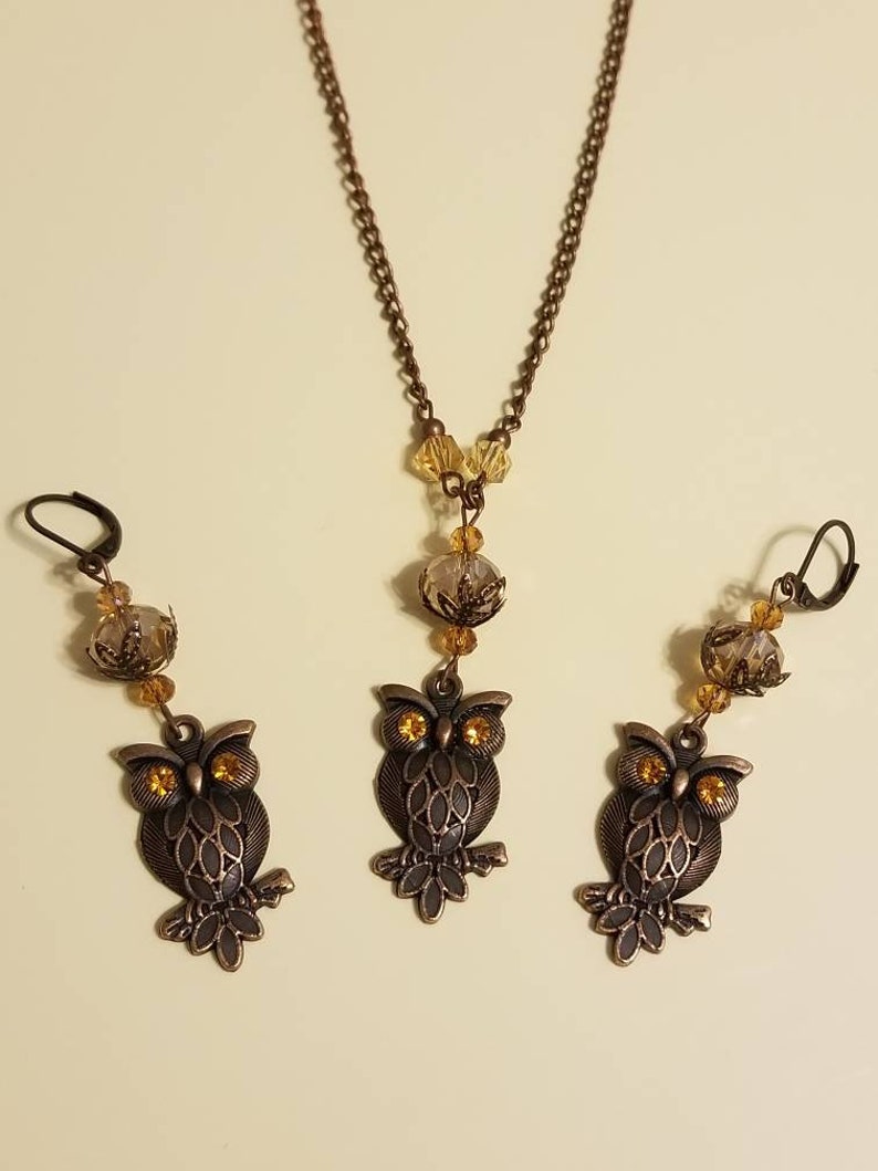May include: A set of owl jewelry featuring a necklace and earrings. The necklace has a bronze chain with a clear crystal pendant and amber-colored beads. The earrings are bronze with amber-colored crystal accents and owl charms.