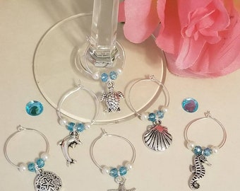 May include: Six silver wine glass charms with blue and white beads. Each charm features a different sea creature: a turtle, a dolphin, a seashell, a starfish, a seahorse, and a sand dollar.
