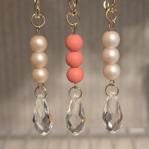 May include: Three gold chain necklaces with different pendant styles. The first necklace has three white pearl beads and a clear crystal teardrop pendant. The second necklace has three coral beads and a clear crystal teardrop pendant. The third necklace has three white pearl beads and a clear crystal teardrop pendant.