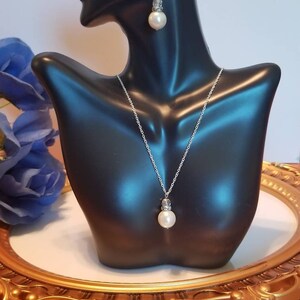 May include: A silver necklace and earrings set with a single large white pearl pendant and matching earrings. The necklace and earrings have a silver chain and a small crystal accent above the pearl.