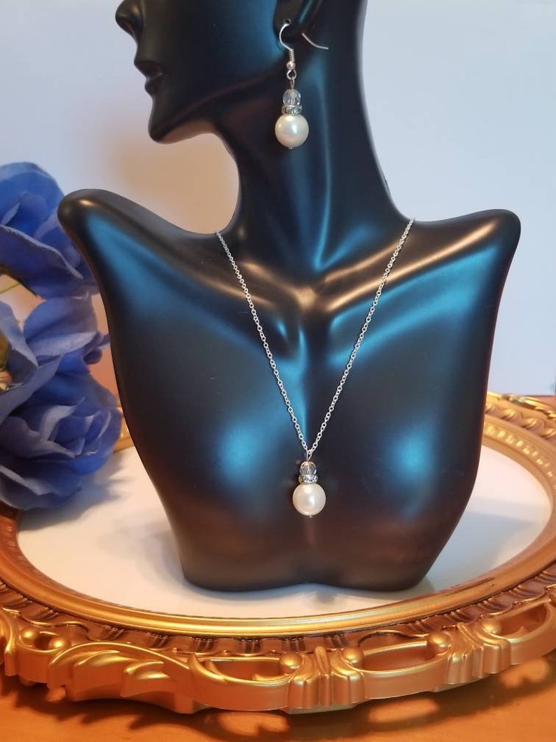 May include: A silver necklace and earrings set with a single large white pearl pendant and matching earrings. The necklace and earrings have a silver chain and a small crystal accent above the pearl.