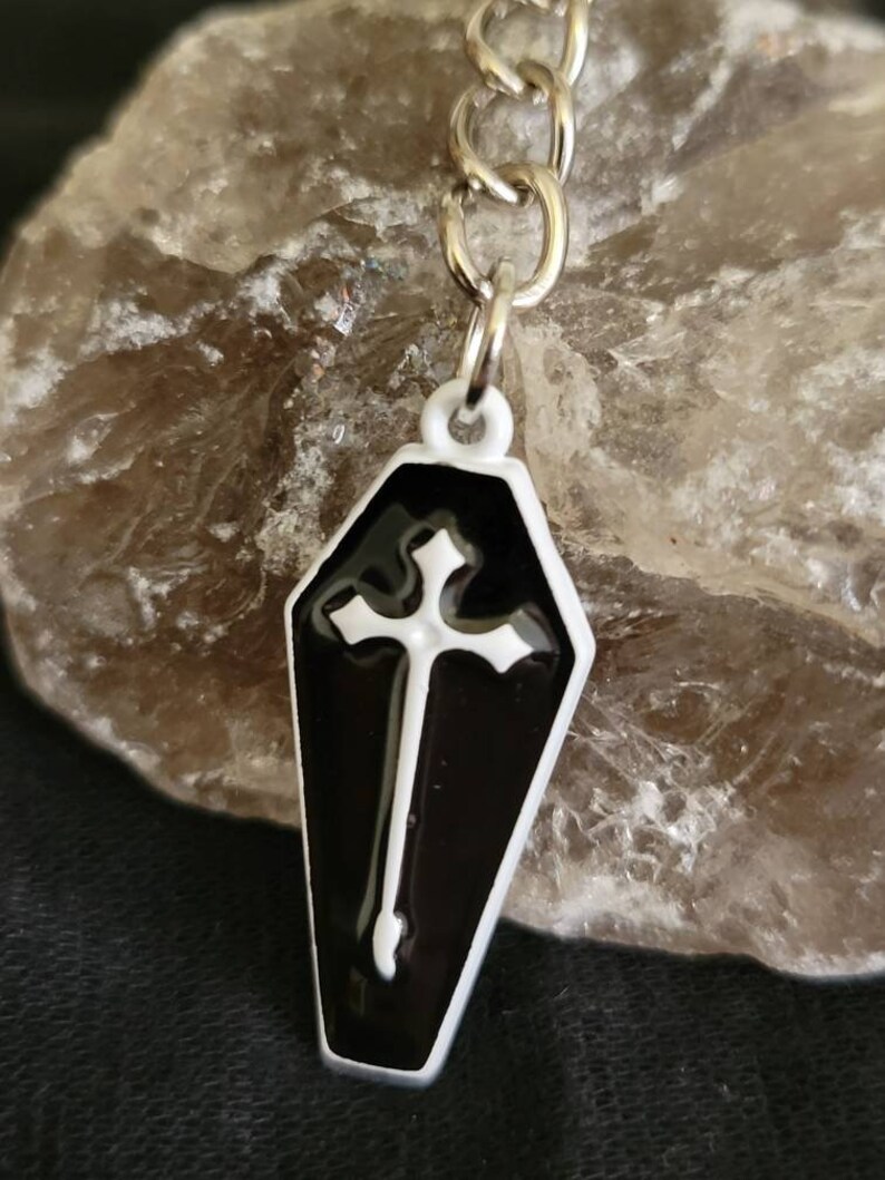 May include: A black and white coffin-shaped keychain with a white cross on the front. The keychain is attached to a silver chain.