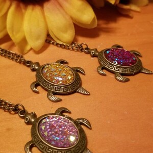 May include: Three turtle-shaped pendant necklaces with bronze-colored metal frames and chains. Each turtle has a different colored, glittery center: gold, purple, and pink. A yellow sunflower is in the background.