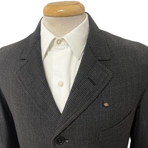 Könnte beinhalten: Ein anthrazitfarbener Blazer mit einem dezenten Karomuster, kombiniert mit einem weißen Hemd mit Kragen. Der Blazer hat einen Zwei-Knopf-Verschluss und ein fallendes Revers. Die Jacke wird auf einer Schaufensterpuppe präsentiert.