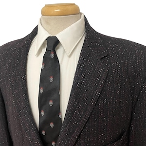 May include: A dark tweed blazer with a white dress shirt and a black tie. The tie features a repeating pattern of a crest with red accents. The blazer has a classic two-button design.