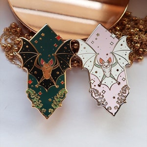 May include: Two enamel pins featuring bats with a star and diamond pattern. The bat on the left is brown and the pin is green. The bat on the right is white and the pin is pink.