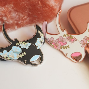 May include: Two enamel pins featuring manta rays with floral designs. One pin is black with blue flowers and the other is white with pink flowers.
