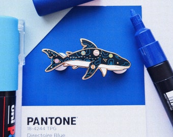 May include: A blue and white enamel pin featuring a shark with a galaxy pattern. The pin is on a blue background with the text "PANTONE 18-4244 TPG Directoire Blue".