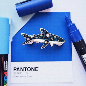 May include: A blue and white enamel pin featuring a shark with a galaxy pattern. The pin is on a blue background with the text "PANTONE 18-4244 TPG Directoire Blue".