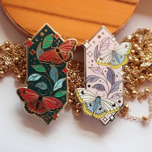 May include: Two decorative enamel pins with butterfly designs. One pin is dark green with red and gold accents, the other is light pink with blue and yellow accents. Both pins have gold-colored borders and are set against a gold chain.