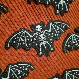 Peut inclure: Un ensemble de patchs thermocollants en feutre noir représentant une chauve-souris squelette avec des étoiles et des croissants de lune blancs.