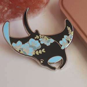 May include: A black enamel pin with a floral design featuring blue flowers and green leaves. The pin is shaped like a manta ray.