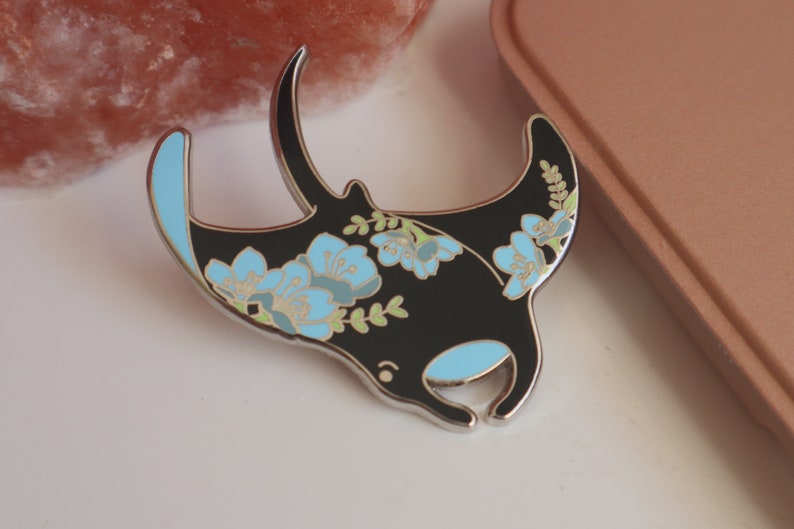 May include: A black enamel pin with a floral design featuring blue flowers and green leaves. The pin is shaped like a manta ray.