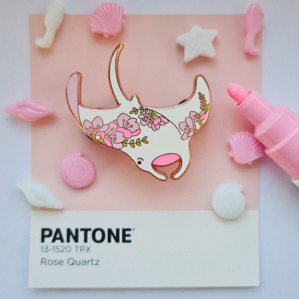 May include: A white enamel pin featuring a pink floral design of a manta ray. The pin is displayed on a pink background with a white card that reads "PANTONE 13-1520 TPX Rose Quartz".