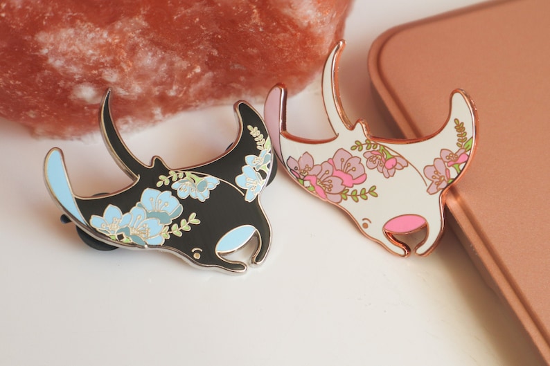 May include: Two enamel pins featuring manta rays with floral designs. One pin is black with blue flowers and the other is white with pink flowers.