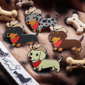 Op de afbeelding: Emaille pins met teckelhonden die cowboyhoeden en rode bandana's dragen. De pins zijn in verschillende kleuren, waaronder zwart, bruin en grijs, met gouden accenten. Hondenkoekjes in de vorm van botten en een halsband zijn ook zichtbaar.