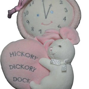 Puede incluir: Un reloj de peluche rosa y blanco con un ratón que sostiene un corazón rosa que dice "Hickory Dickory Dock".