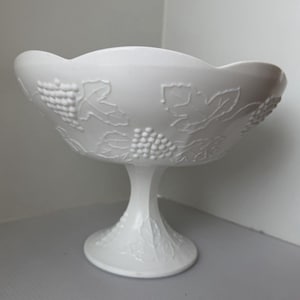 Puede incluir: Una compotera blanca con pie y borde festoneado. El cuenco está decorado con diseños en relieve de uvas y hojas. La base del pedestal también está decorada con motivos de hojas. Una pieza decorativa para servir fruta o exhibir objetos.