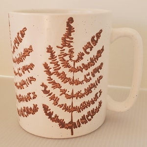 May include: White ceramic mug with a brown fern leaf design. The mug has a speckled finish and a large handle. The fern design is detailed and covers a large portion of the mug's surface. The mug is suitable for hot or cold beverages.