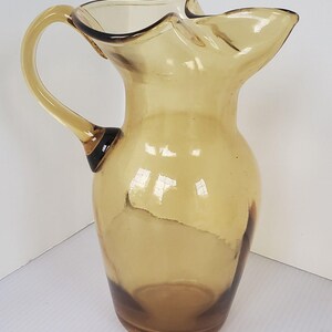 May include: A vintage amber glass pitcher with a ruffled rim and a curved handle. The pitcher has a classic design and is made of transparent glass. The handle is attached to the body of the pitcher with a dark brown accent.