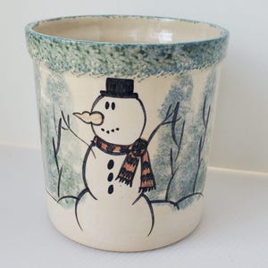 Pode incluir: Um porta-utensílios de cerâmica com um desenho de boneco de neve pintado à mão. O boneco de neve usa um chapéu preto e um cachecol com listras laranja e pretas. O porta-utensílios tem uma borda verde e azul e um fundo de cena de inverno.