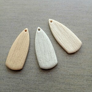 May include: Three teardrop-shaped pendants in varying shades of beige and off-white. Each pendant has a small hole at the top for stringing and a textured surface resembling wood grain. The pendants are displayed on a neutral gray surface.