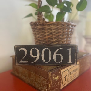 Może przedstawiać: Czarny drewniany znak z białymi malowanymi cyframi "29061" na czerwonej powierzchni.