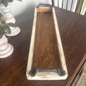 May include: A rectangular wooden trough with a distressed white painted frame and a natural wood interior. The trough has black metal handles on each end. This decorative piece is suitable for home decor.