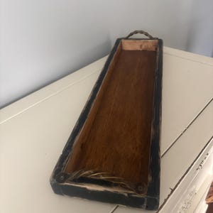 Op de afbeelding: Een rechthoekige houten dienblad met een donker gebeitst interieur en zwart geschilderde buitenkant. De lade heeft een geweven handvat en decoratieve metalen accenten. De verweerde afwerking geeft het een rustieke, boerderij-esthetiek.