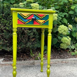 May include: A small, lime green end table with a single drawer. The drawer front is painted with a colorful geometric pattern in shades of red, yellow, blue, and green. The table has four turned legs.