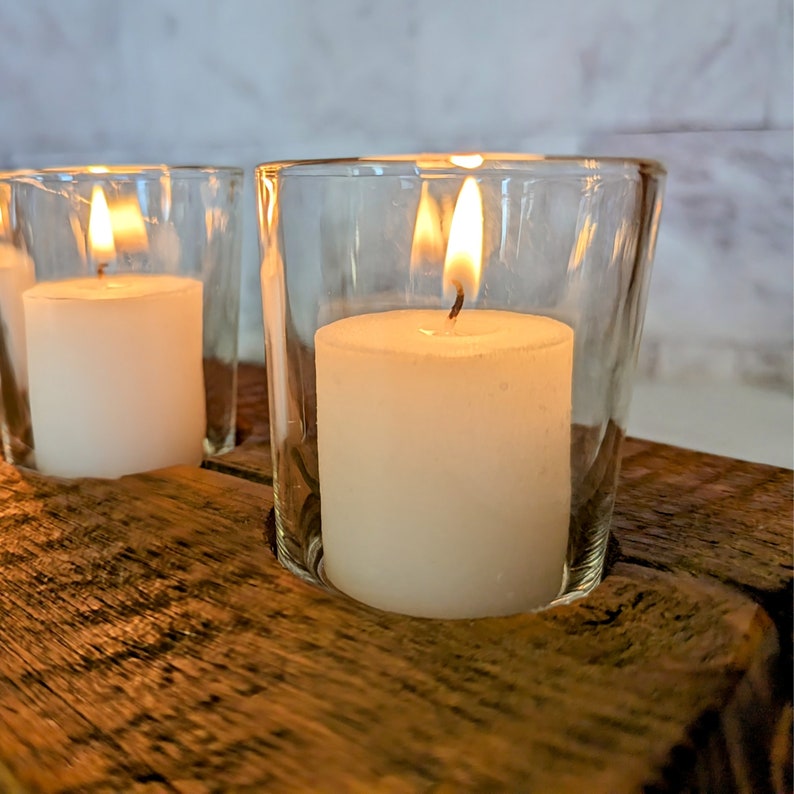 May include: Close-up of lit white candles in clear glass votive holders. The candles are set on a rustic, weathered wooden surface. The flames are bright and the glass reflects the light.