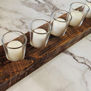 May include: A rustic wooden candle holder with five clear glass votive holders, each containing a white pillar candle. The wood has a rich, dark brown color with visible grain and knots. The candles are unlit, and the background is a marble-patterned surface.