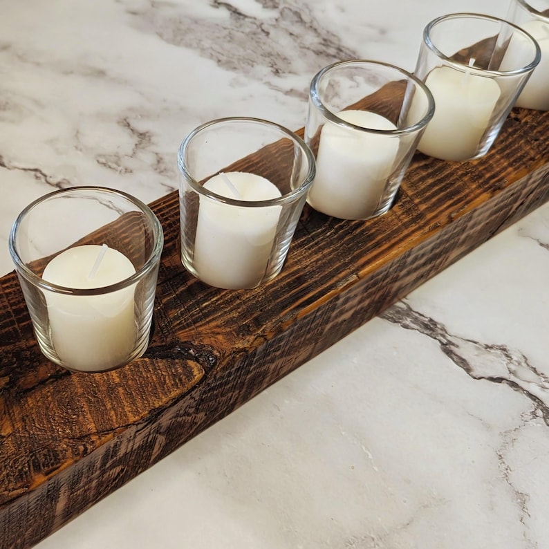 May include: A rustic wooden candle holder with five clear glass votive holders, each containing a white pillar candle. The wood has a rich, dark brown color with visible grain and knots. The candles are unlit, and the background is a marble-patterned surface.